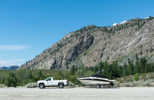 A profile shot of the 2025 GMC Canyon AT4X towing a small boat trailer on a paved road. The focus is on the truck's stance and the secure connection between the truck and the trailer. The background shows a scenic lake and mountains.