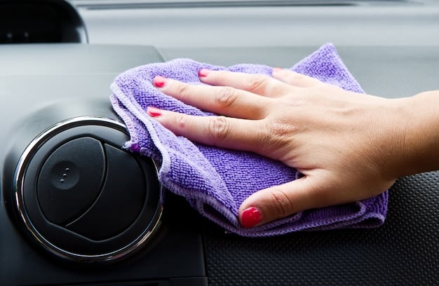 A hand wiping down a car dashboard with a microfiber cloth, with bottles of cleaning products visible in the background.